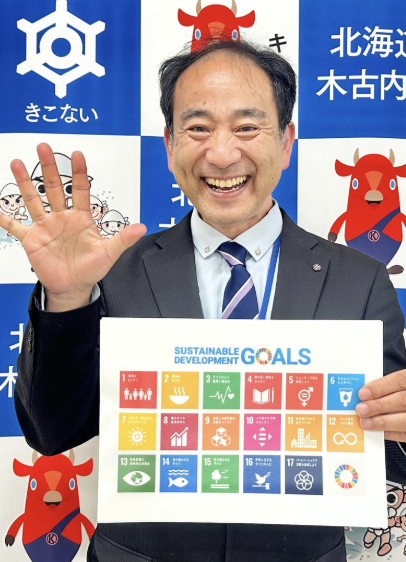 木古内町の背景の前で、SDGsのパネルを手に笑顔で手を振るモリモリ先生